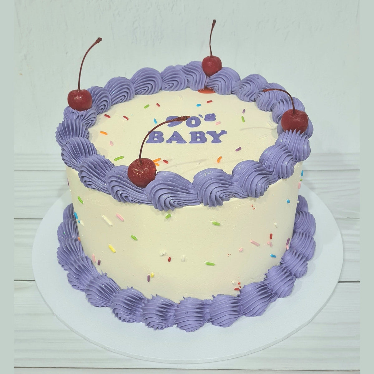 Cake with purple frosting, cherries, and '90's Baby' text on a white background