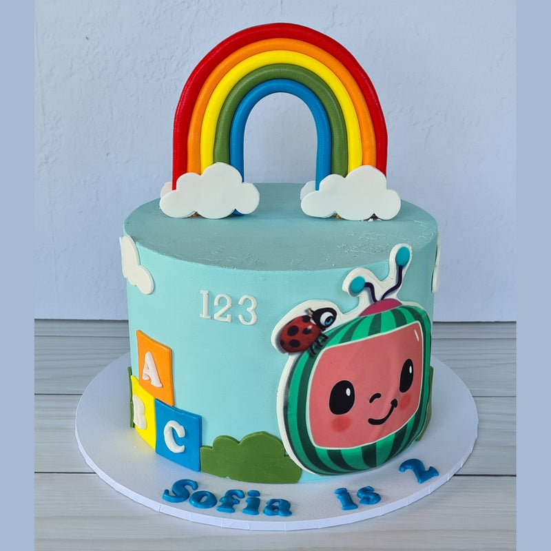 Children's birthday cake with a rainbow, watermelon character, and colorful elements on a light blue background.