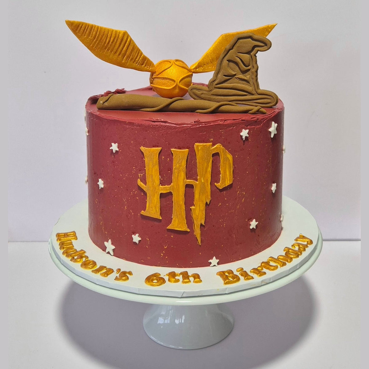 Red birthday cake with gold 'HP' letters and a golden snitch on top, on a white stand.
