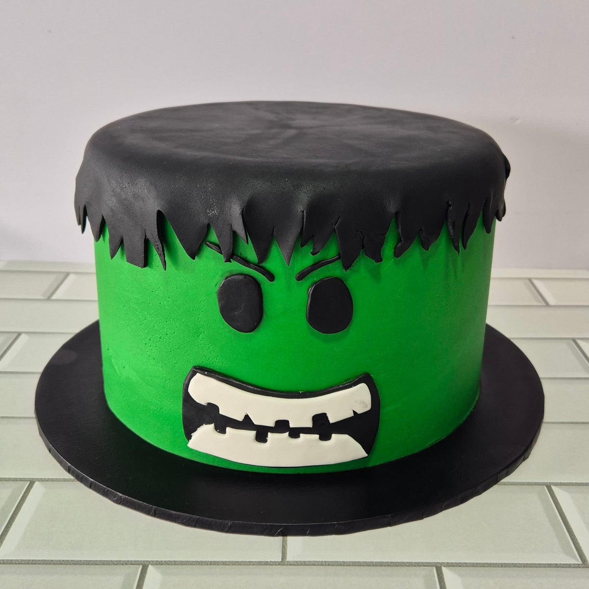 Cake designed to resemble the Hulk with green body, black top, and white mouth on a tiled floor.