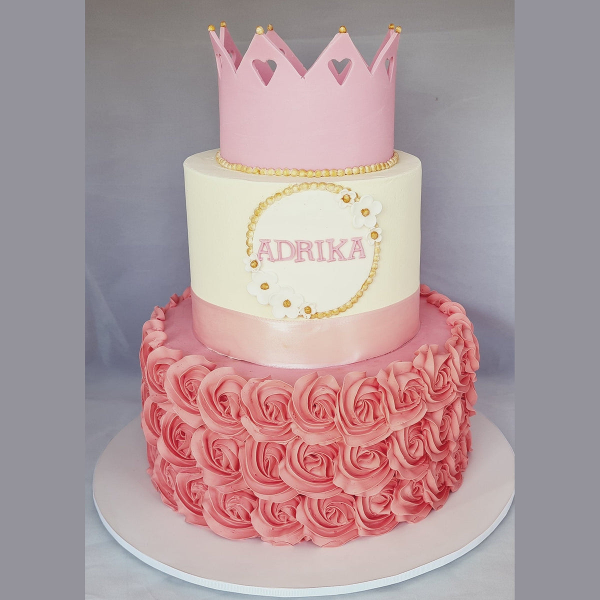 Two-tiered cake with a pink crown and 'ADRIKA' on a gray background
