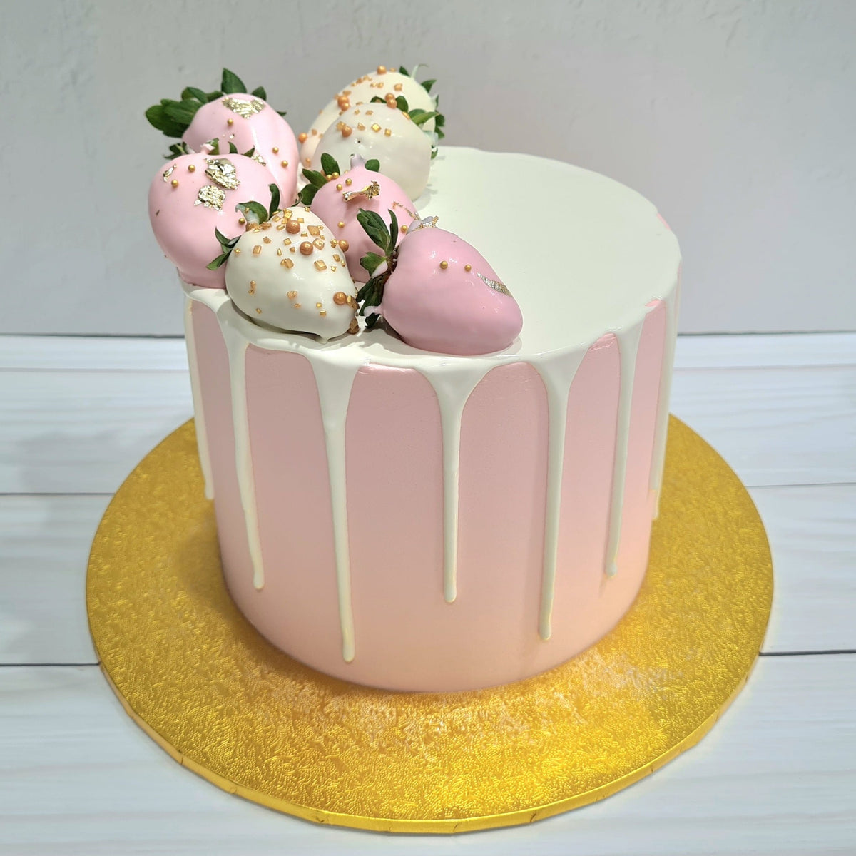 Pink cake with white drizzle and decorated strawberries on a gold cake stand.