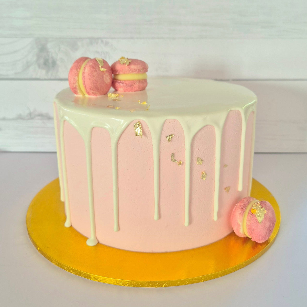Pink cake with white drizzle and macarons on a gold cake stand against a white background